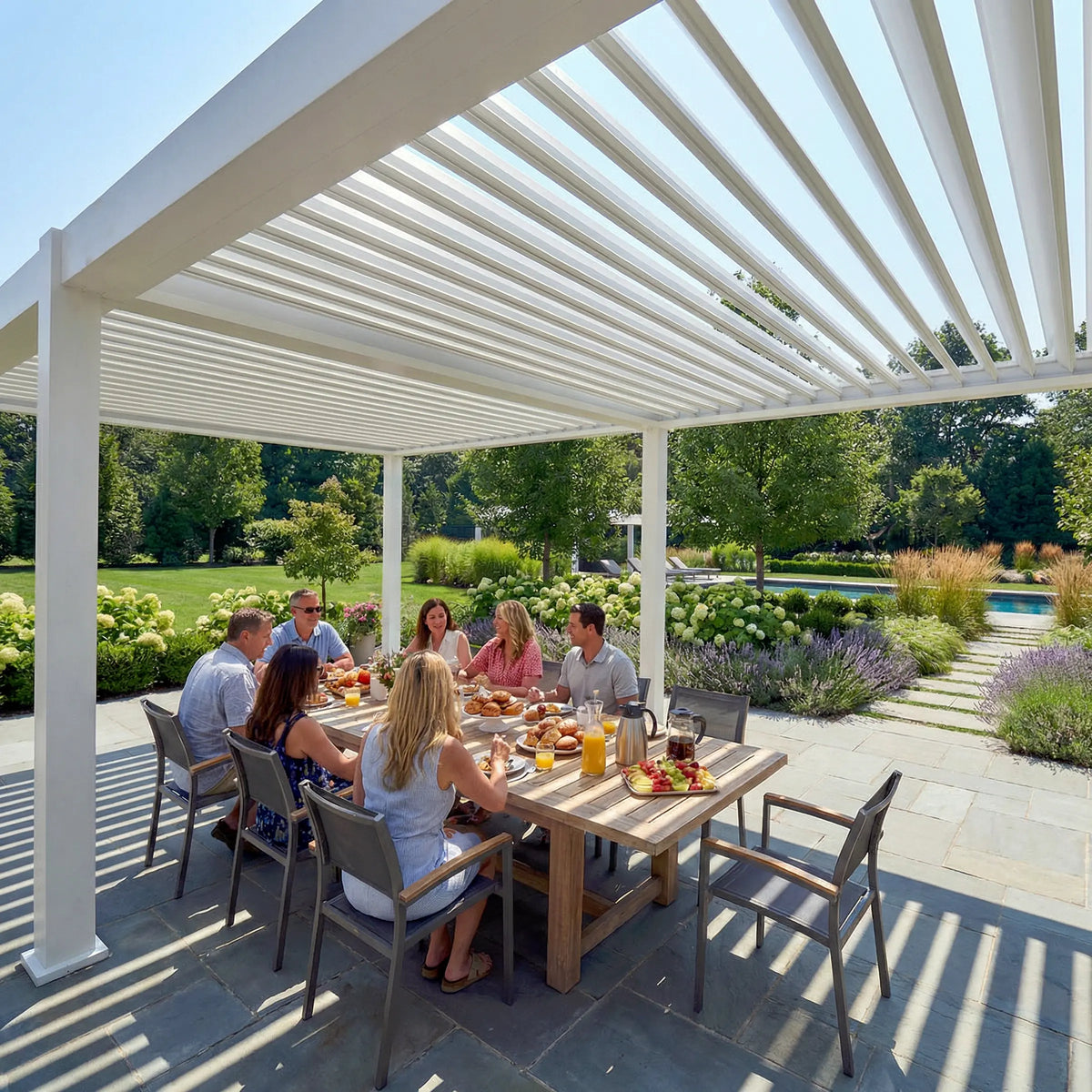 People dining outdoors under a white pergola on a sunny patio surrounded by landscaped gardens.