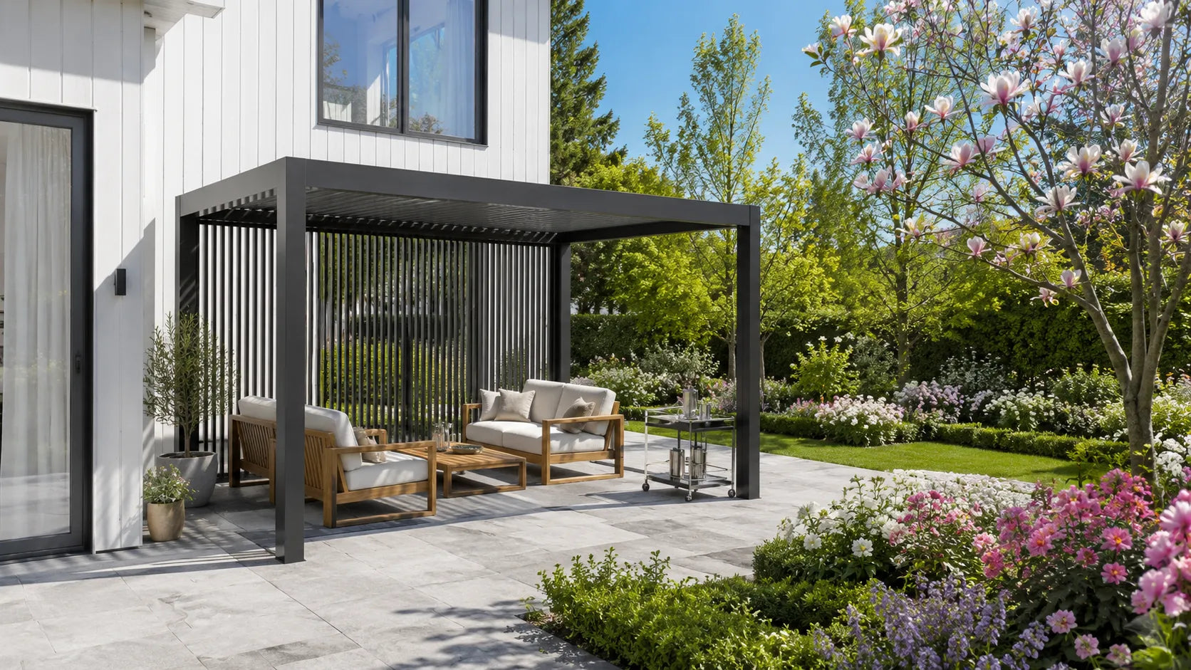 Black modern pergola with a slatted privacy wall sheltering a patio seating area next to a lush, colorful garden.